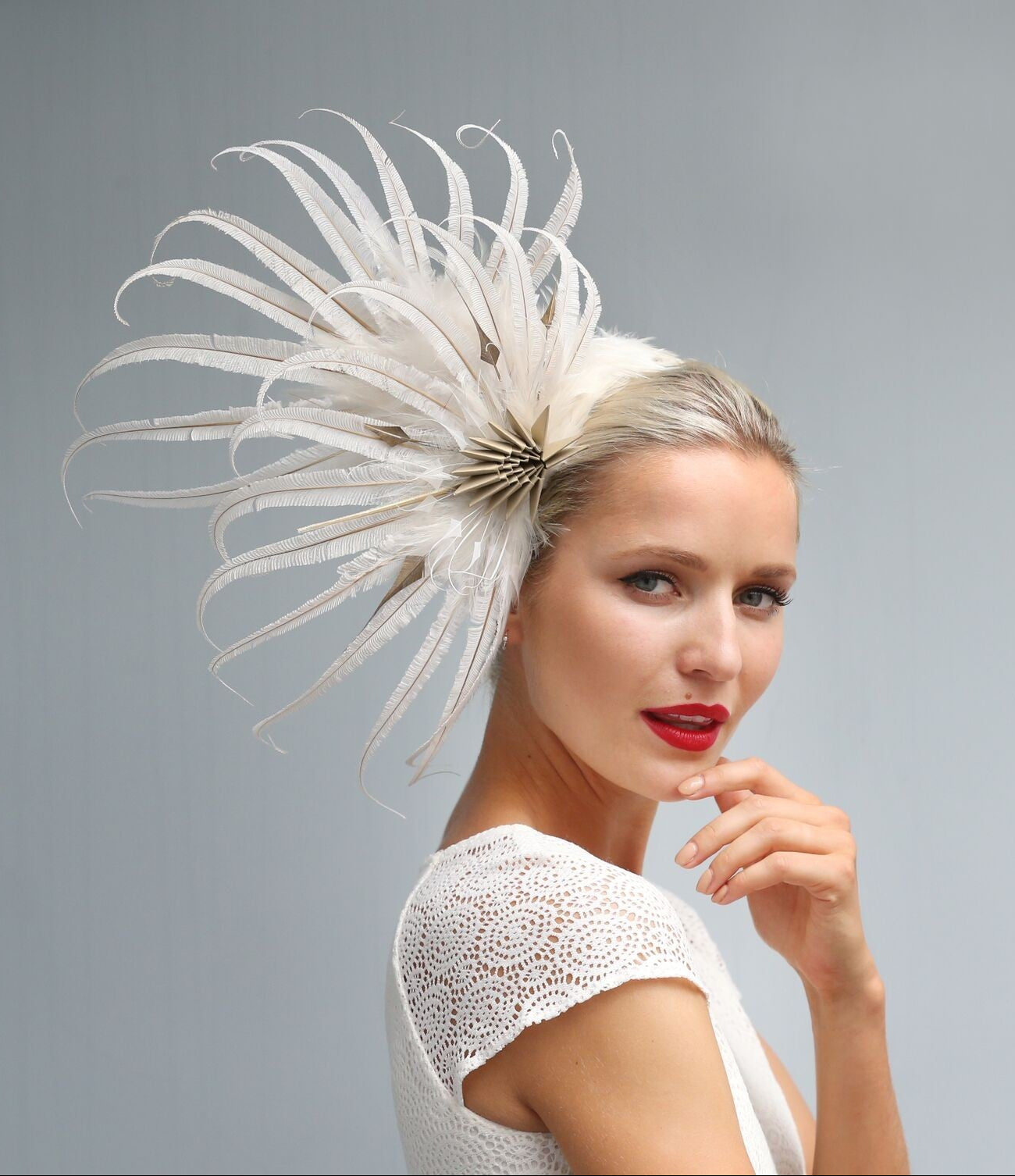 A woman in a white lace top models a dramatic white feather fascinator headpiece against a solid gray studio background.