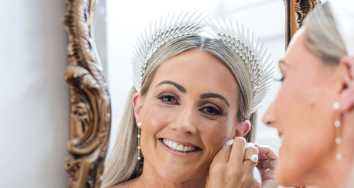 A smiling woman in a white bridal veil looks into an ornate gold mirror while applying makeup. Her reflection shows her wearing a matching tiara, and another woman is partially visible in the foreground.